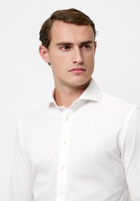 Camicia bianca con bottoni, collo appuntito, realizzata in tessuto liscio, con una fila di bottoni chiari e una silhouette aderente.