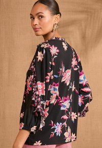 Svart blommig blus med puffärmar, som har livfulla mönster av rosa och blå blommor, samt en volangdetalj vid halslinningen.