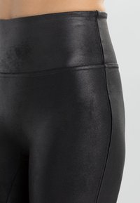 Høytlivs sorte leggings laget av elastisk, skinnende stoff med glatt tekstur, som har en enkel søm langs midjen.