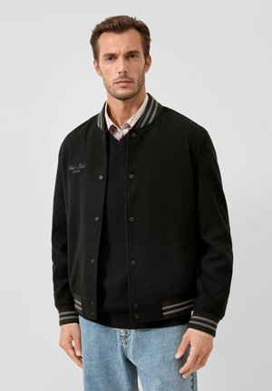 Mann trägt schwarze Collegejacke mit gestreiften Bündchen und Kragen, schwarzen Pullover, rosa Hemd und hellblaue Jeans vor einfarbigem Hintergrund.