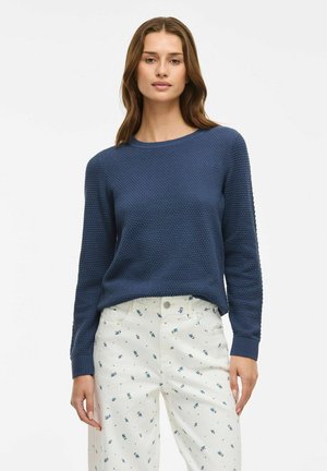 Marineblauwe gebreide trui met ronde hals en lange mouwen, gecombineerd met witte broek met een blauw bloemenpatroon.