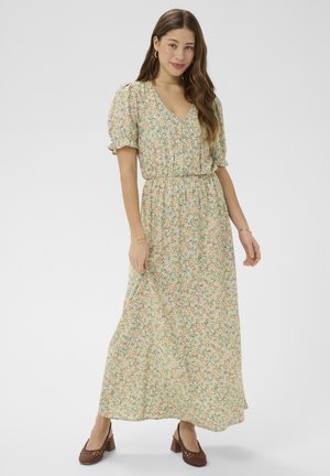 Vrouw die een lichtgroene maxi-jurk met bloemenprint en pofmouwen draagt, samen met bruine gevlochten schoenen met blokhak, staand tegen een effen achtergrond.