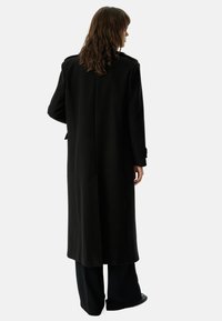 Long black coat made of a soft fabric, featuring a tailored fit, slight sheen, and button accents on the sleeves.