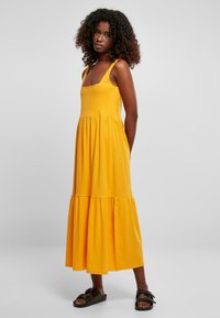 Geel katoenen maxi-jurk met brede schouderbanden, aansluitend bodice en gelaagde ruches aan de onderkant. Gekleed met zwarte sandalen, staande tegen een effen achtergrond.