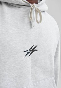 Jasnoszara bluza z kapturem o miękkiej fakturze. Ma czarny graficzny logo na piersi oraz beżowy sznurek przy kapturze.
