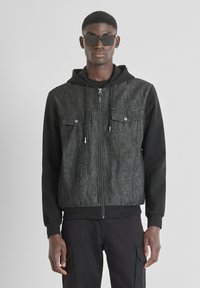 Chaqueta de mezclilla negra con un frente texturizado gris, dos bolsillos en el pecho, capucha y cierre con cremallera. El modelo lleva gafas de sol y pantalones oscuros.
