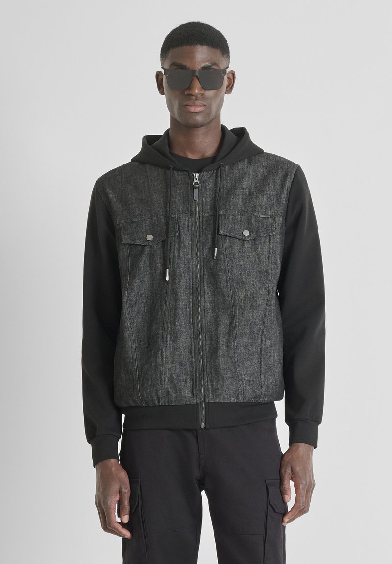 Chaqueta de mezclilla negra con un frente texturizado gris, dos bolsillos en el pecho, capucha y cierre con cremallera. El modelo lleva gafas de sol y pantalones oscuros.