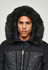Giacca a vento nera con imbottitura a forma di pallone, caratterizzata da un motivo geometrico testurizzato, zip dorata e un ampio cappuccio in pelliccia sintetica con ciocche scure.