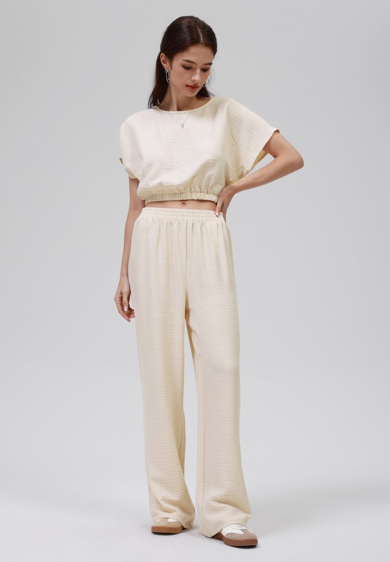 Cremefarvet loungsæt bestående af en cropped top med struktureret overflade og elastisk talje, kombineret med løstsiddende, høj talje bukser.
