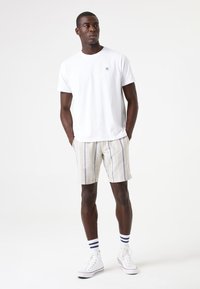 Camiseta blanca de algodón con un pequeño logo, combinada con pantalones cortos de rayas claras en beige y azul, y zapatillas deportivas blancas de corte bajo con acentos en azul.