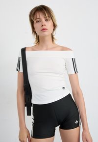Top branco de manguita curta e com ombros à mostra, com detalhes em preto nas três riscas; combinado com calções pretos com riscas brancas laterais e logótipo.