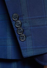 Niebieska tkanina w kratę z czterema czarnymi guzikami z tekstem, teksturowanym splotem i kontrastującymi jasnoniebieskimi akcentami. Zbliżenie na rękaw eleganckiego garnituru.