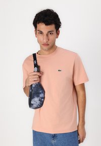 Tricou din bumbac roz deschis cu un logo mic, asortat cu o geantă crossbody de culoare albastru închis, cu tie-dye, având un fermoar și o curea.