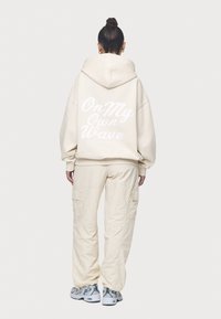 Överdimensionerad beige hoodie med vit text "On My Own Wave" på baksidan, ihop med matchande beige cargobyxor och grå sneakers.