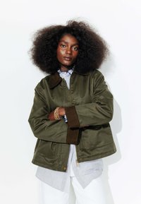 Veste vert olive avec un col marron, comportant une fermeture éclair et deux poches avant. Superposée à une chemise rayée et un pantalon blanc.