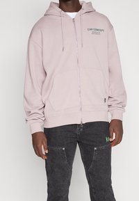 Sudadera con capucha rosa claro con cremallera frontal, bolsillo delantero, capucha con cordón y texto en el lado izquierdo del pecho. Tejido suave y corte relajado, combinada con jeans oscuros.