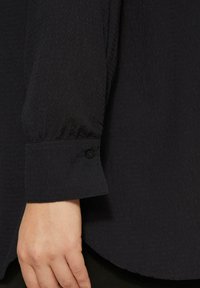 Zwarte textuur met lange mouwen shirtmanchet met knoopdetail. De stof lijkt lichtgewicht en heeft een subtiel kreukpatroon. Ronde zoom zichtbaar.