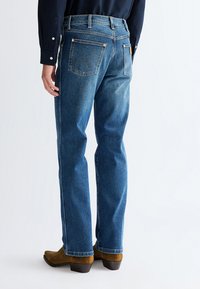 Mann trägt blaue Jeans mit geradem Bein und braune Wildlederstiefel, steht mit dem Rücken zur Kamera vor einem einfarbigen Hintergrund.