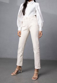 Hvid skjortebluse med plisserede ærmer, parret med off-white lige ben jeans og nude hæle med stropper på en grå overflade.