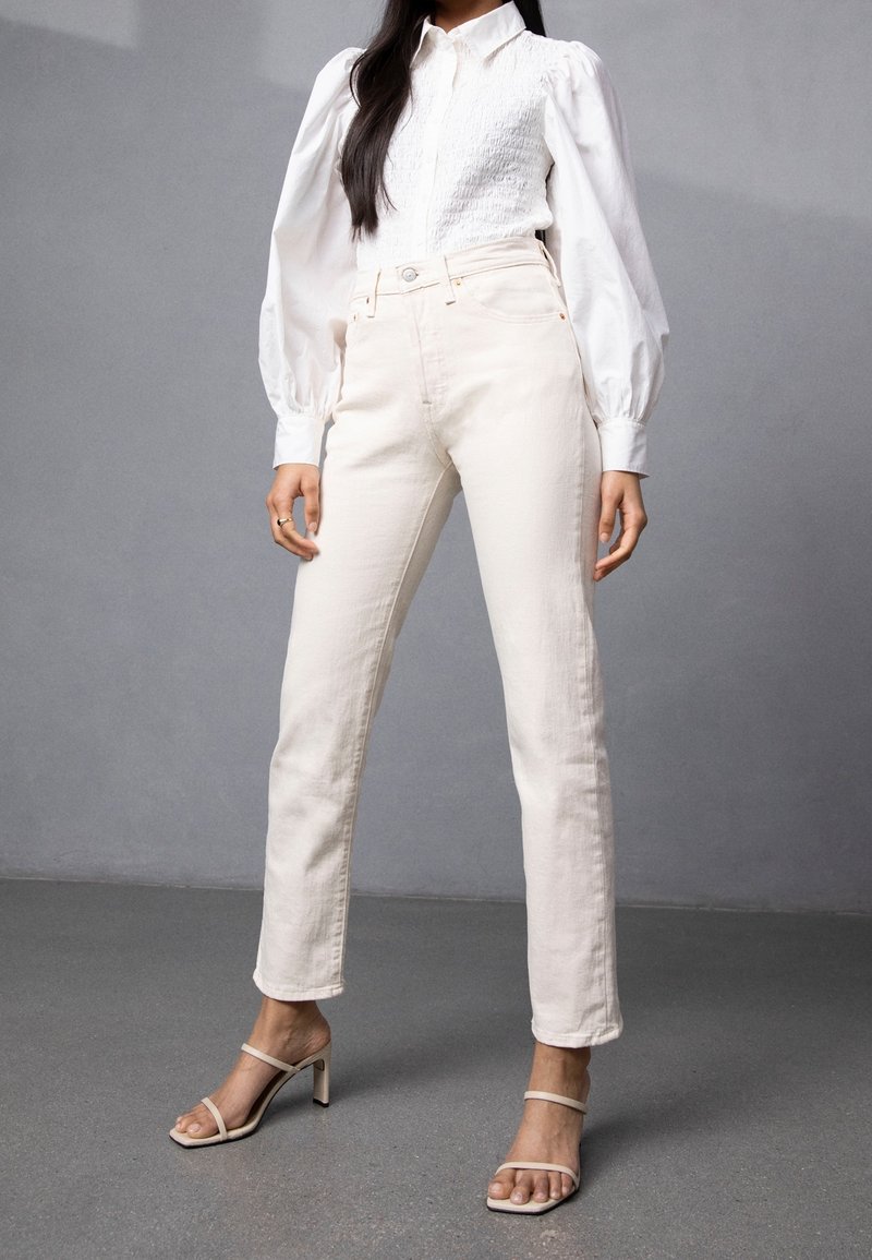 Hvid skjortebluse med plisserede ærmer, parret med off-white lige ben jeans og nude hæle med stropper på en grå overflade.