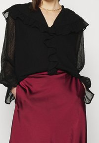 Blusa nera con volant e maniche lunghe trasparenti, abbinata a una gonna in satin bordeaux. La blusa presenta un scollo a V e chiusure con bottoni.