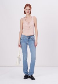 Lys rosa ribbet tanktop med blondekant og knapper, kombinert med lyseblå tettsittende jeans; modellen holder en hvit håndveske og har på seg svarte sko.