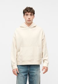 Νέος άνδρας με σγουρά μαλλιά, ντυμένος με εκρού φούτερ και μπλε τζιν, στέκεται μπροστά σε απλό φωτεινό φόντο.