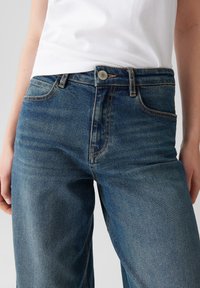 Close-up van een persoon in blauwe spijkerbroek en een wit shirt, met de nadruk op de taille, zakken en knoopdetails.