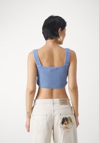 Top court en tricot bleu avec de larges bretelles ; texture côtelée. Porte avec un jean crème présentant un design brodé et une étiquette à l'arrière.