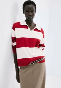 Donna che indossa un maglione a righe rosse e bianche con colletto, gonna beige e cintura a catena nera e oro, in piedi davanti a uno sfondo bianco.