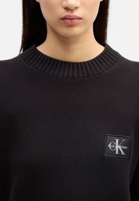 Črn pleten pulover z rebrastim ovratnikom in kvadratno nalepko z logotipom "Calvin Klein" ter stiliziranim "K" v beli barvi.