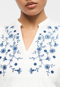 Gros plan sur un vêtement blanc avec un col en V orné de broderies florales bleues sur la poitrine et les épaules.