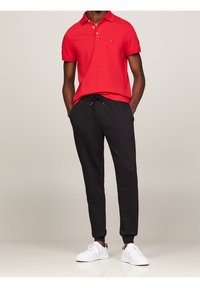 Röd polo med tre knappar, kombinerad med svarta joggers med dragsko och vita sneakers, vilket visar en avslappnad passform.