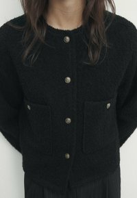 Svart ullcardigan med en fluffig textur, med två framfickor och fem knappar i guldton.