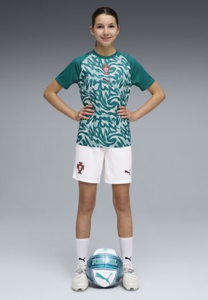 Joven jugadora de fútbol con las manos en las caderas, vistiendo el uniforme del equipo de Portugal y tacos, con un balón de fútbol a sus pies.