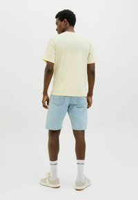 Geel T-shirt met korte mouwen, lichtblauwe denim shorts, witte sokken met zwart logo, en beige sneakers. Achteraanzicht tegen een effen achtergrond.