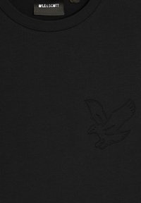 Sort bomulds t-shirt med et tekstureret ørne-emblem i samme farve, rund hals og en glat overflade. Mærkets etiket er synlig ved halsudskæringen.