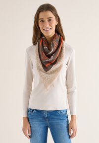 Femme souriante portant un haut à manches longues beige clair, un jean bleu et une grande écharpe marron et beige avec des accents rouges autour du cou.