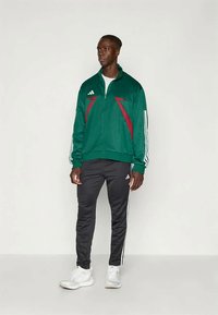 Πράσινη και κόκκινη αθλητική ζακέτα με λευκές ρίγες στα μανίκια, συνδυασμένη με μαύρο παντελόνι που έχει λευκές ρίγες στο πλάι, φορεμένη με λευκά αθλητικά παπούτσια.