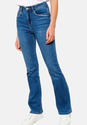 Hoch taillierte blaue Jeans mit ausgestelltem Bein, fünf Taschen, vorn mit Knopfverschluss und gestickten Details. Getragen mit weißen Sneakers.