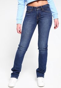Jeans de denim azul ajustados con una cintura de altura media, que presentan un diseño clásico de cinco bolsillos y un desvanecimiento mínimo. Llevados con zapatillas blancas.
