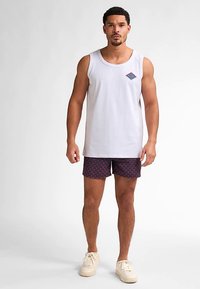 Vitt ärmlöst linne med en liten mångfärgad diamantlogotyp, kombinerat med marinblå shorts med geometriskt mönster och krämfärgade sneakers.