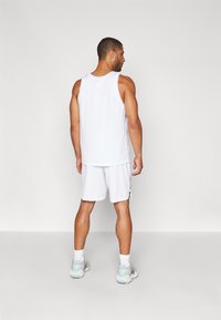 Homem de costas, vestindo uma camisola desportiva sem mangas branca, calções brancos, meias brancas e sapatilhas brancas, em frente a um fundo liso.