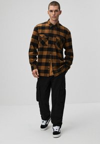 Brązowa i czarna w kratkę flanelowa koszula z dwoma kieszeniami na piersi, połączona z czarnymi spodniami cargo i czarno-białymi trampkami.