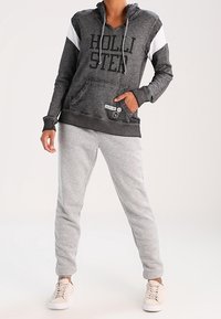 Šedá mikina s kapucí s černými a bílými doplňky a nápisem "HOLLISTER"; kombinovaná se světle šedými tepláky a krémovými teniskami.