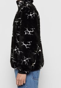 Personne portant une veste polaire noire avec des motifs blancs en forme d'éclairs, debout de profil, associée à un jean en denim bleu.