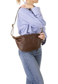 Borsa a spalla in pelle marrone con forma curvata, tracolla regolabile, chiusura a zip e texture liscia. Indossata sopra una camicia a righe blu.