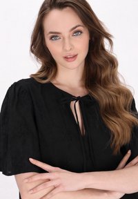 Černý top s vázáním u krku, krátkými nabíranými rukávy a texturovaným květinovým vzorem. Modelka má vlnité vlasy a má zkřížené paže.