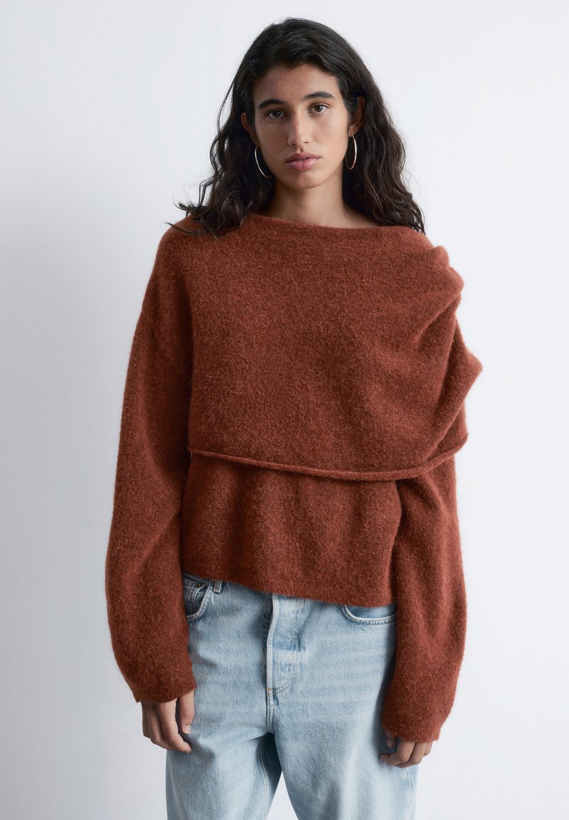 Textured rust-colored pullover sweater with an oversized design, wide neckline, and long sleeves, layered at the waist over a fitted, smooth top.