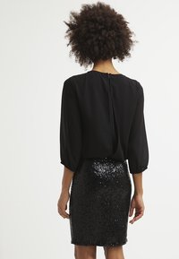 Nainen, jolla on kiharat hiukset, pukeutuneena mustaan pitkähihaiseen paitaan ja polvenpituiseen mustaan strassihameeseen, seisoo selkä kameraan päin.